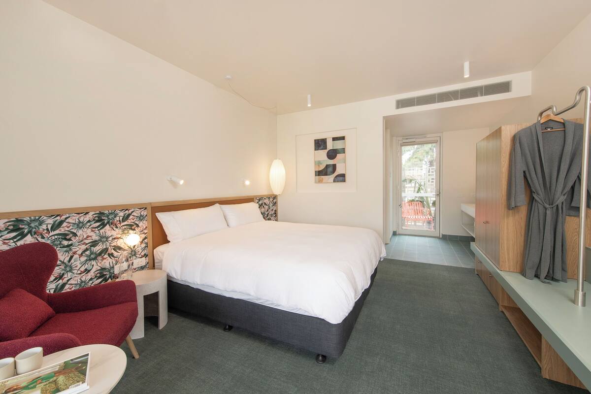 A hotel room with a bed, a red armchair, a small table with a lamp, and a wardrobe.