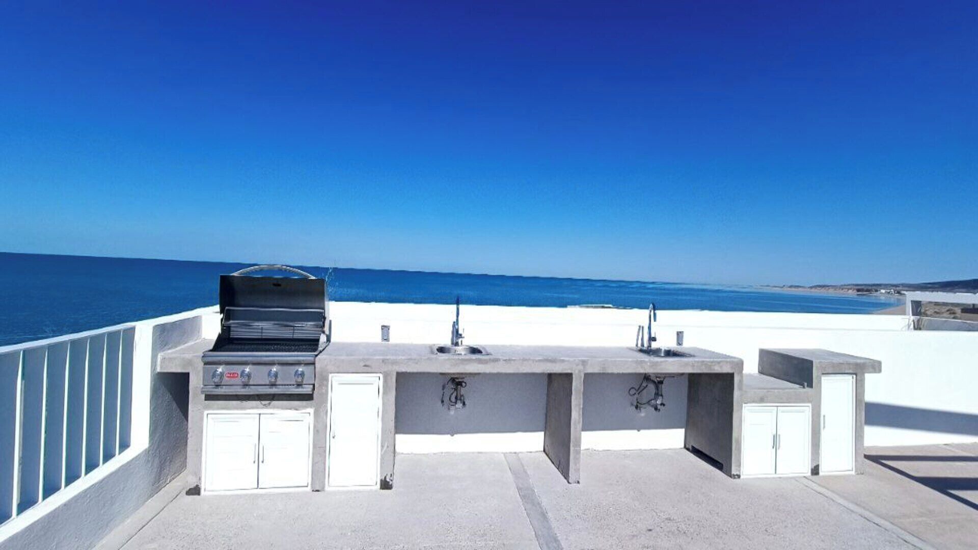 An outdoor kitchen with a grill, two sinks, and storage cabinets, overlooking a beach.