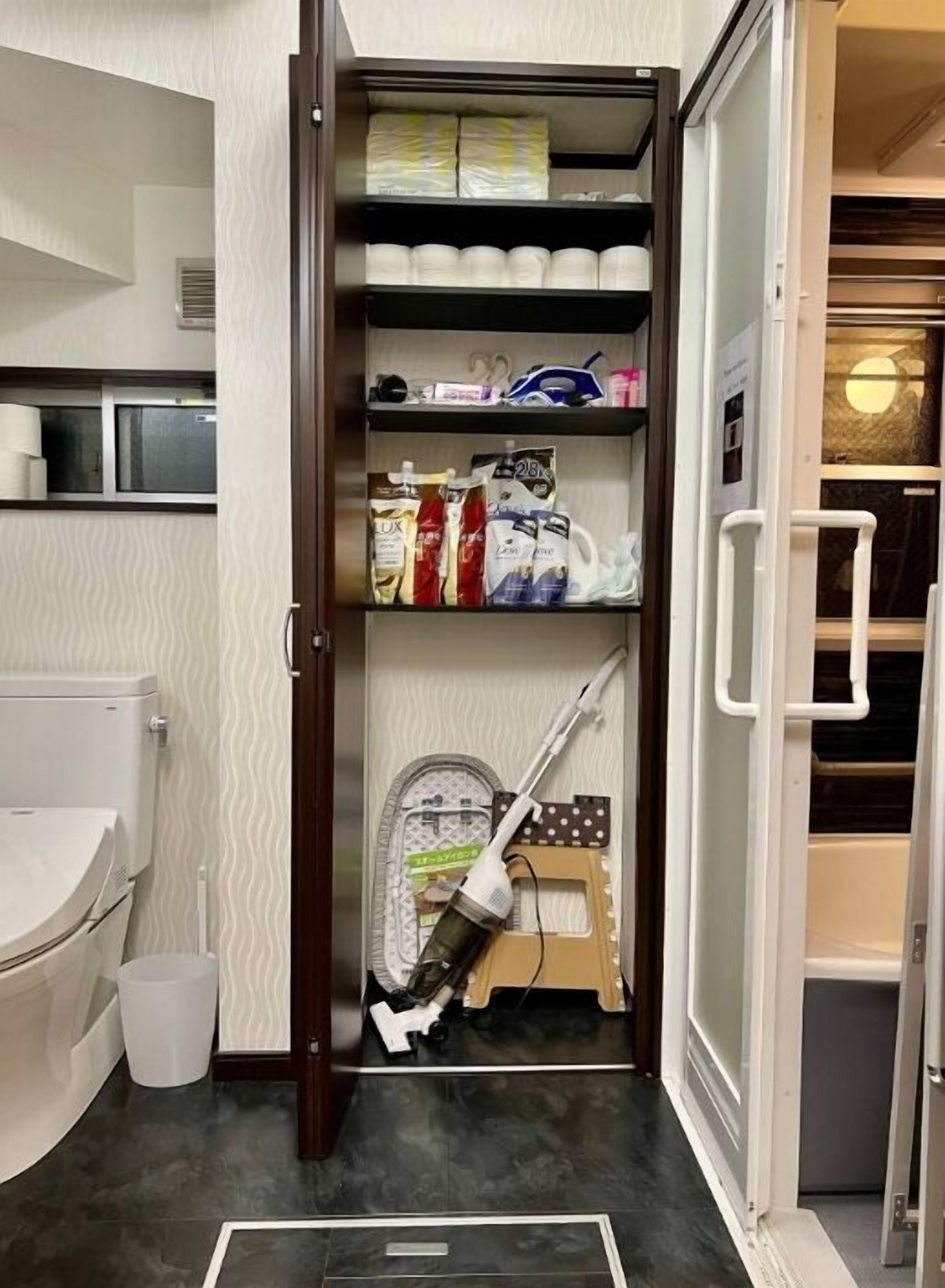 A compact bathroom with a toilet, sink, and a shelf with toiletries and cleaning supplies.