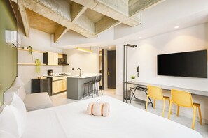 A modern hotel room with a bed, a desk with two chairs, a kitchen area, and a TV mounted on the wall. - Firenze Lofts (Medellín)