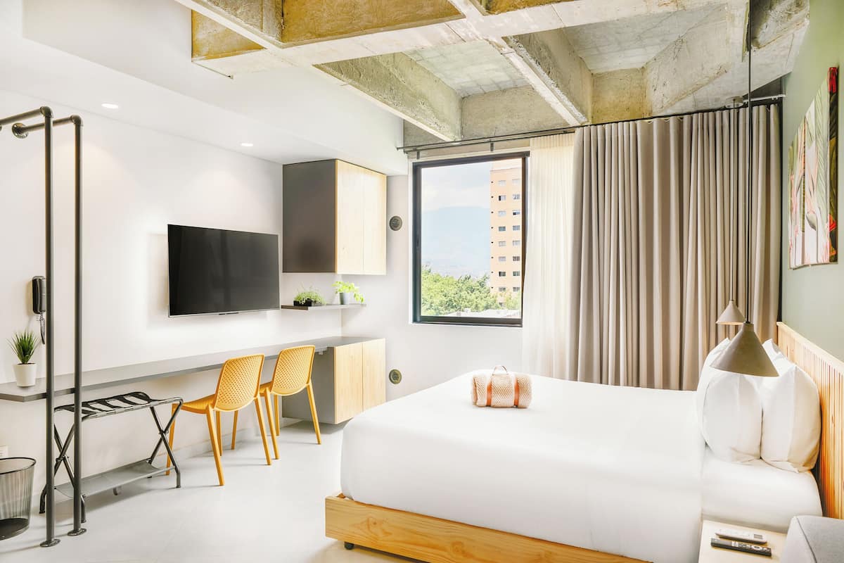 Una habitación de hotel moderna con una cama grande, un escritorio con dos sillas, un televisor montado en la pared y una ventana con cortinas.