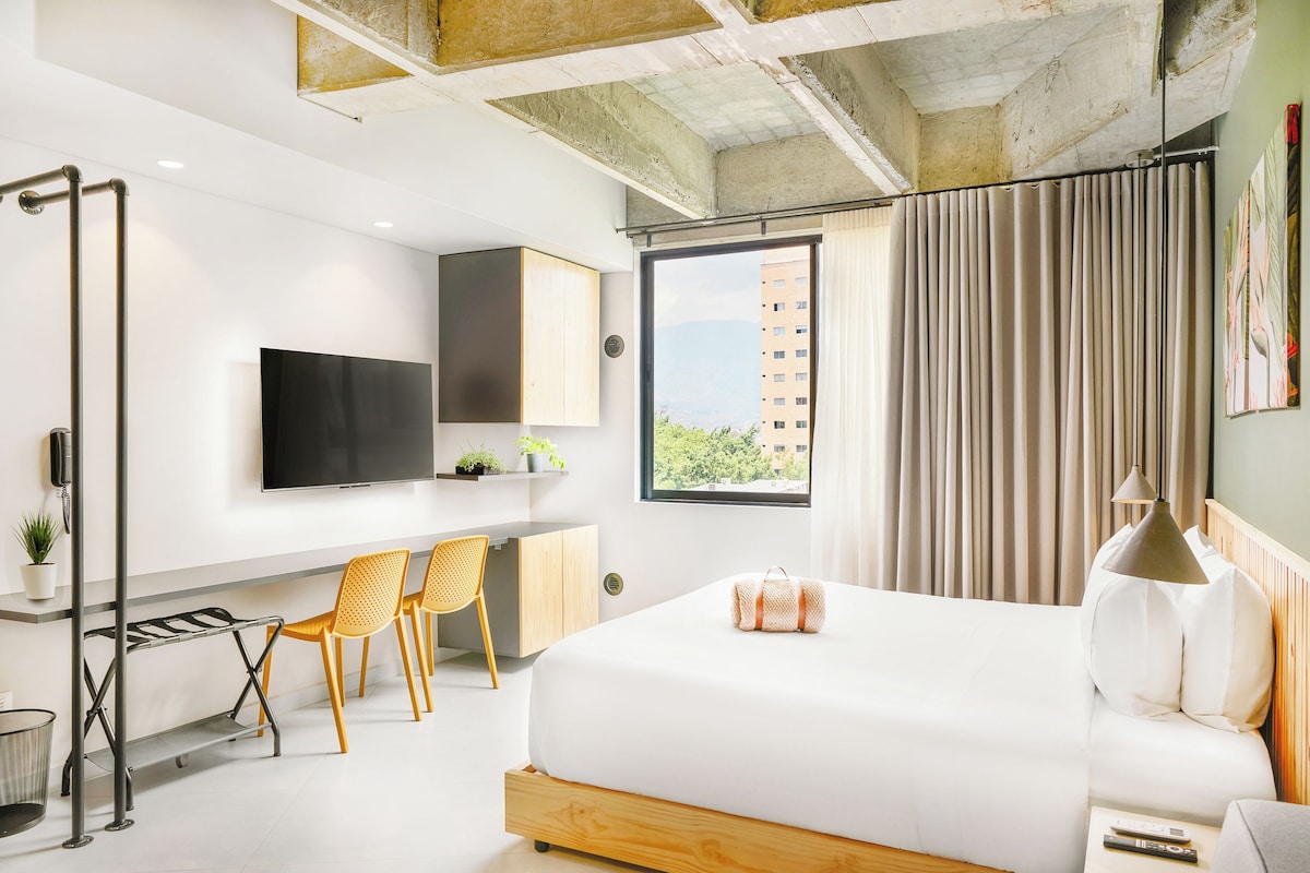 A modern hotel room with a large bed, a desk with two chairs, a TV mounted on the wall, and a window with curtains.