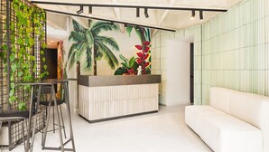 A reception area with a modern design, featuring a white reception desk, a white sofa, and a mural of tropical plants. - Firenze Lofts (Medellín)