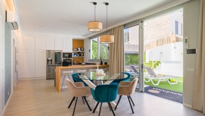 A modern kitchen with a central island, a dining area with a glass table and chairs, and a view of a garden through large sliding doors.