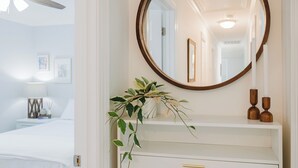 Een slaapkamer met een bed, nachtkastje met lamp, spiegel en een plant op een dressoir.