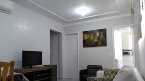 A living room with a television on a wooden stand, a sofa, and a painting on the wall. - Round trip - LA12-Bertioga, SESC, Riviera, Vista Linda, Indaiá (Jardim Rafael Bertioga)