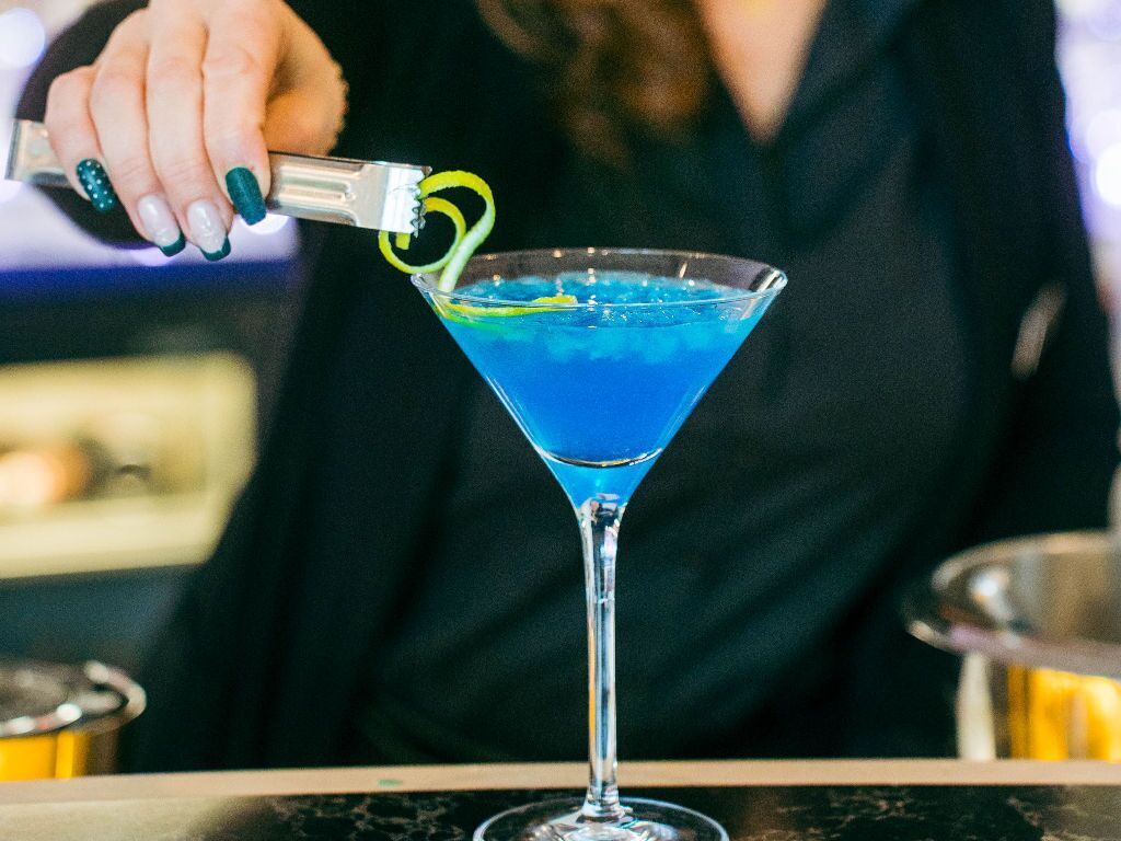 A bartender pouring a blue cocktail into a martini glass with a lemon twist garnish.