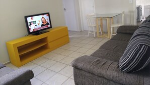 Ein Wohnzimmer mit einem Fernseher auf einem gelben Schrank, einem grauen Sofa, einem weißen Esstisch mit Stühlen und einer weißen Tür.