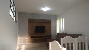 Ein Wohnzimmer mit einem an einer Holzwand montierten Flachbildfernseher, einem braunen Sofa und einem weißen Esstisch mit Stühlen.