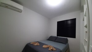 Ein kleines Zimmer mit einem Einzelbett, einer dunkelblauen Tagesdecke, zwei Kissen und einer an der Wand montierten Klimaanlage.