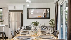 Comedor con mesa de madera dispuesta para seis personas, rodeada de sillas, con cocina y un espejo grande en la pared.