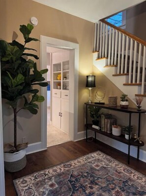 A hallway with a wooden floor, a patterned rug, a potted plant, a console table with decorative items, and a staircase with wooden railings.