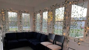 Habitació acollidora amb un sofà seccional de color fosc i una sola cadira, decorada amb plantes penjants i llums de cadena.