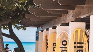 Varias tablas de surf con el texto 'Mahalo Hawaii', ubicadas bajo una estructura de madera en la playa.