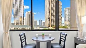 Una habitación con una mesa redonda, dos sillas, una planta pequeña en maceta y vistas de edificios altos a través de grandes ventanales.