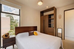 A bedroom with a bed, a nightstand, a wardrobe, and a window with a view of trees. - Beautiful 2 Bedroom Apartment (Tulum)