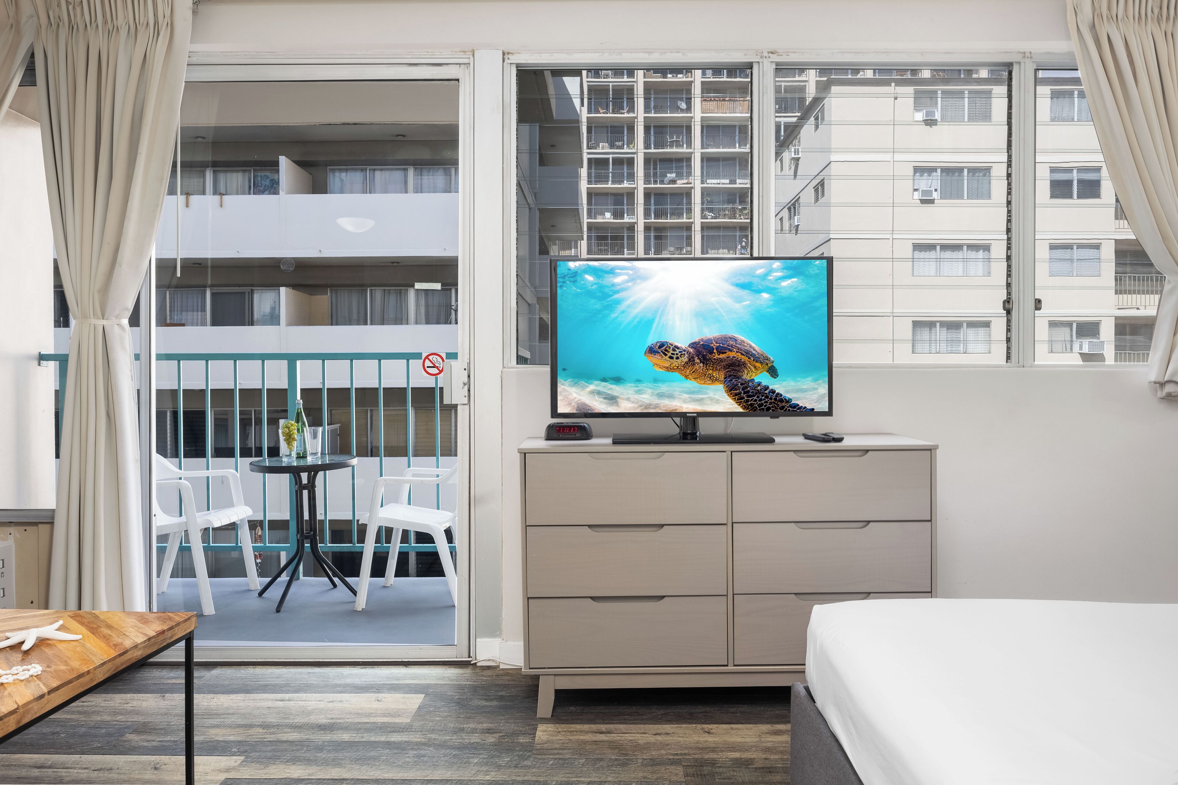 Une chambre d’hôtel avec un lit, un téléviseur diffusant une image de tortue, une petite table avec une plante, et un balcon avec des chaises et une rambarde.