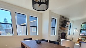 Ein Essbereich mit Holztisch und Bänken, großen Fenstern, einem Steinkamin und einer modernen Leuchte.