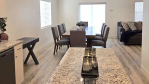 A modern living room with a dining area, featuring a kitchen island, dark-toned furniture, and a large window providing natural light.