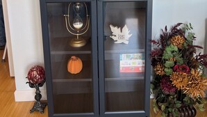 Un mueble con puerta de vidrio y estantes que contiene objetos decorativos como una calabaza blanca, una ampolla de vidrio y una cruz de madera.
