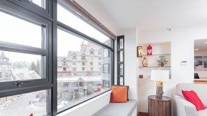 A modern living room with a large window, a sofa, a round wooden side table, and a small shelf with decorative items. - Moscato-Ski-In/Ski-Out (A Condo at Carleton Lodge) (Whistler)