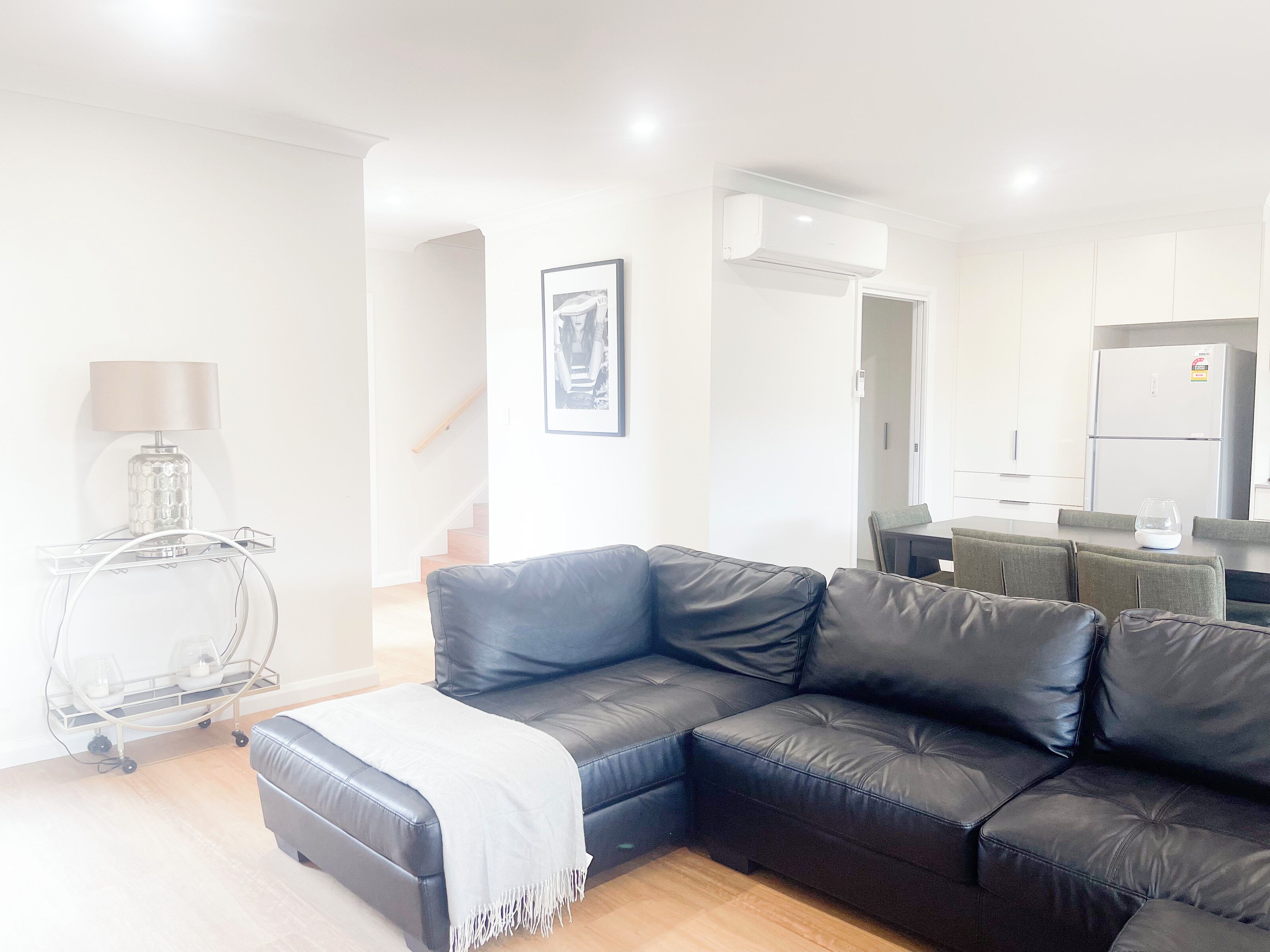 A modern living room with a black leather sofa, a small table with a lamp, and a dining area with a table and chairs.