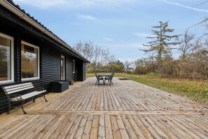 Une terrasse en bois avec un banc et une table, un bâtiment doté de grandes fenêtres et un paysage naturel avec des arbres.
