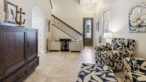 A spacious living area with a dark wooden cabinet, a staircase, and a floral-patterned sofa. - Coastal Retreat – Near the Beach w/ Private Hot Tub & Spacious Backyard Oasis (Indian Harbour Beach)