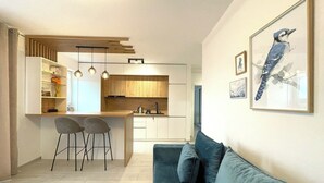 Een moderne keuken met houten kasten, een centraal eiland en hanglampen. In de woonkamer staat een blauwe bank en een houten salontafel.