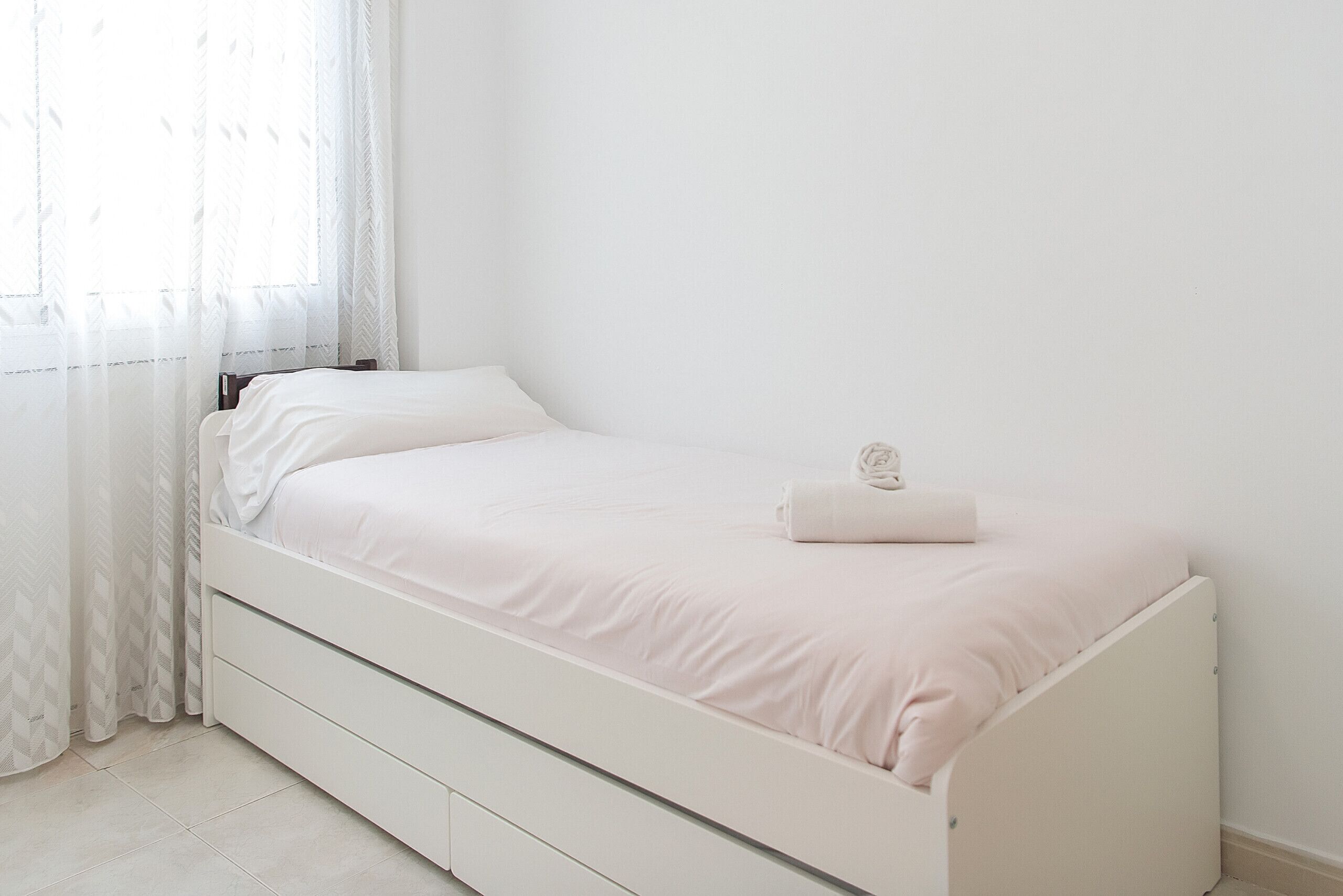 Una cama blanca con un solo cojín y una toalla enrollada, una mesita de noche blanca y una ventana con cortinas traslúcidas.
