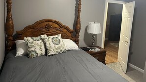 Une chambre à coucher avec un lit à baldaquin, deux coussins à motifs, une table de chevet avec une lampe et une porte donnant sur une autre pièce.