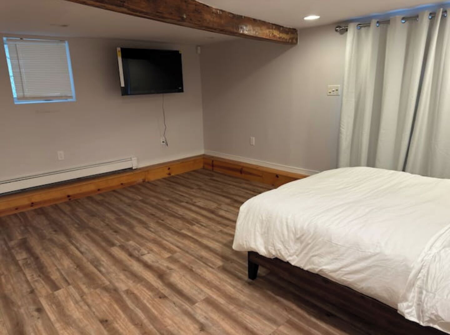 Un dormitorio con piso de madera, una cama, un televisor montado en la pared y un ventanal con persianas.