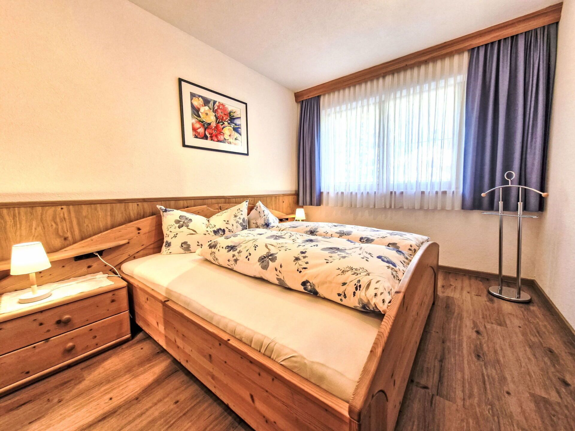 Un lit en bois avec une literie à motifs floraux, une table de chevet avec une lampe, une commode en bois et une fenêtre avec des rideaux.