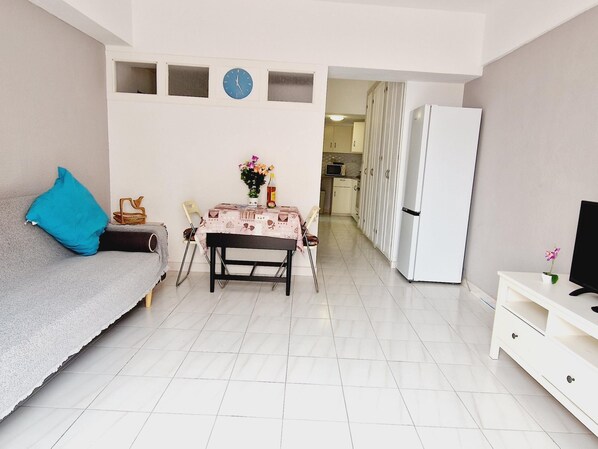Una habitación pequeña y luminosa con sofá, una mesa de comedor con sillas, un refrigerador y un televisor sobre un mueble.