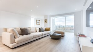 Ein geräumiges Wohnzimmer mit einer großen Couch, einem runden Couchtisch und Blick auf eine Stadtkulisse durch große Fenster.