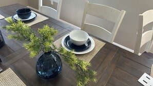 Una mesa de comedor con un florero de follaje, sillas blancas y platos apilados.