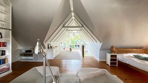 Un'ampia camera da letto mansardata con soffitto alto, pavimento in legno, una grande finestra e un angolo lettura accogliente con una lampada.