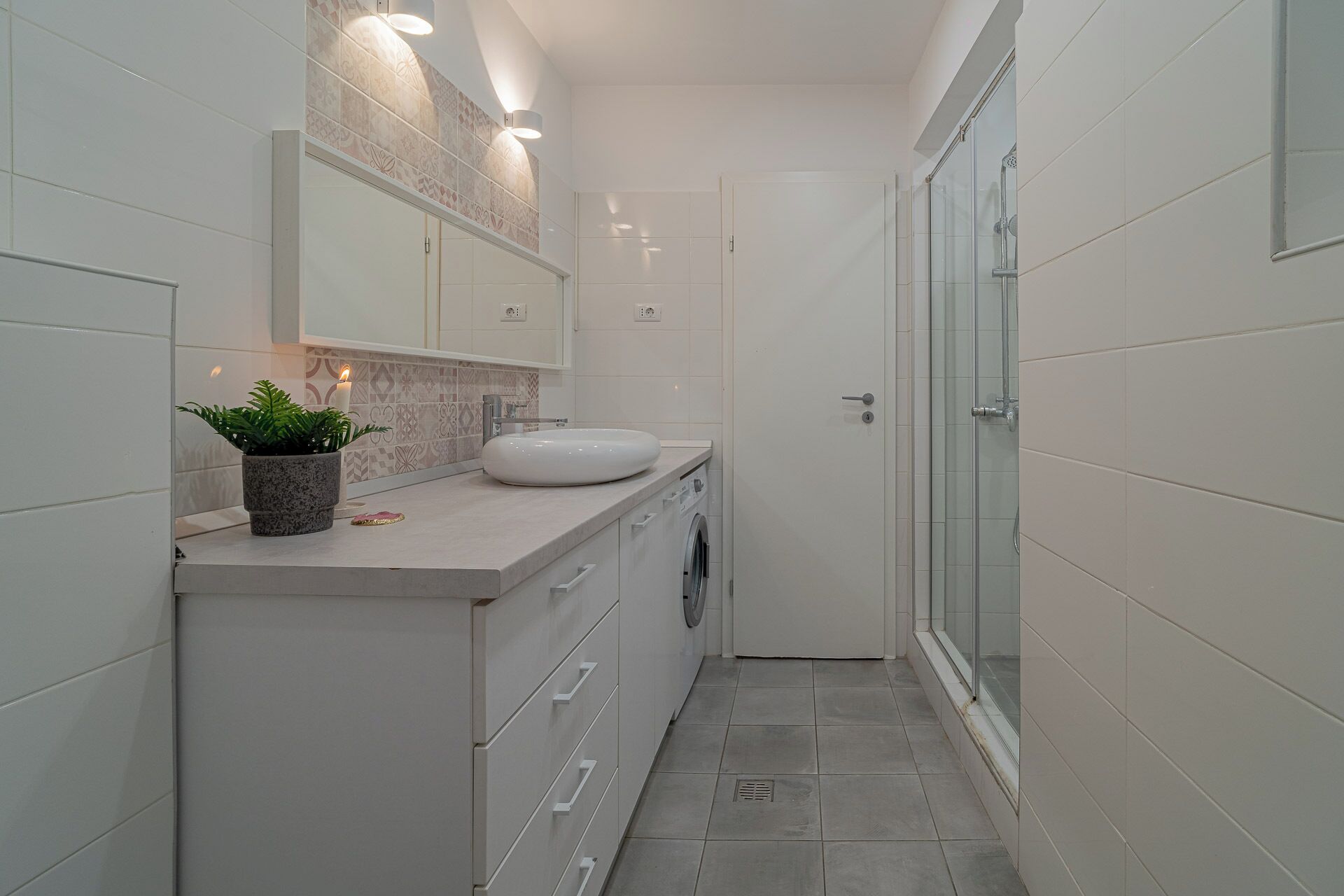 A narrow bathroom with a white sink, a potted plant, and a glass shower enclosure.