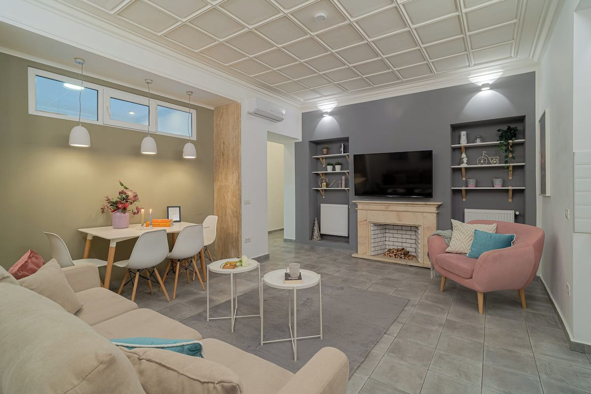 Un salon moderne comprenant un canapé, un fauteuil rose, une table à manger avec des chaises, une cheminée et un téléviseur fixé au mur.