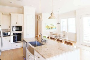 A kitchen with white cabinets, stainless steel appliances, a marble countertop, pendant lighting, and a dining area with a table and chairs.