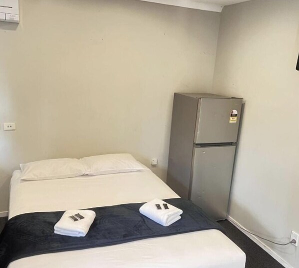 A single bed with white and dark bedding, a small refrigerator, and a power outlet on the wall. - Dash motel (Papakura)