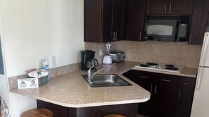 A compact kitchen with a sink, a coffee maker, a toaster, and a microwave. There are two wooden stools and a sign on the counter. - Studio Apartment- Steps from the beach! (Deerfield Beach)