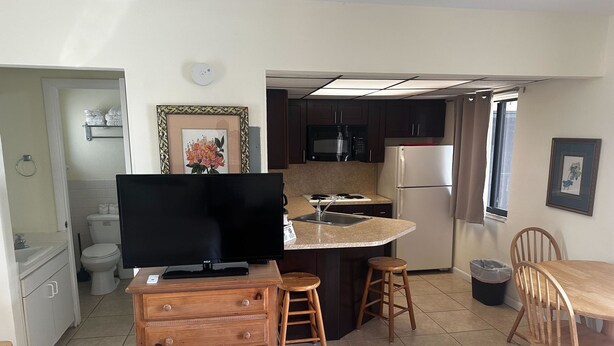 A compact kitchen with a TV, wooden bar stool, and a dining table with chairs. - Studio Apartment- Steps from the beach! (Deerfield Beach)
