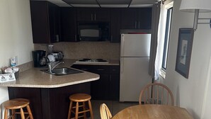 A compact kitchen with dark cabinets, a microwave, a stove, and a refrigerator. There is a wooden dining table with four chairs and a small wooden stool. - Studio Apartment- Steps from the beach! (Deerfield Beach)