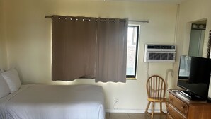 A bedroom with a bed, a ceiling fan, an air conditioning unit, a television, a wooden dresser, and a window with curtains. - Studio Apartment- Steps from the beach! (Deerfield Beach)