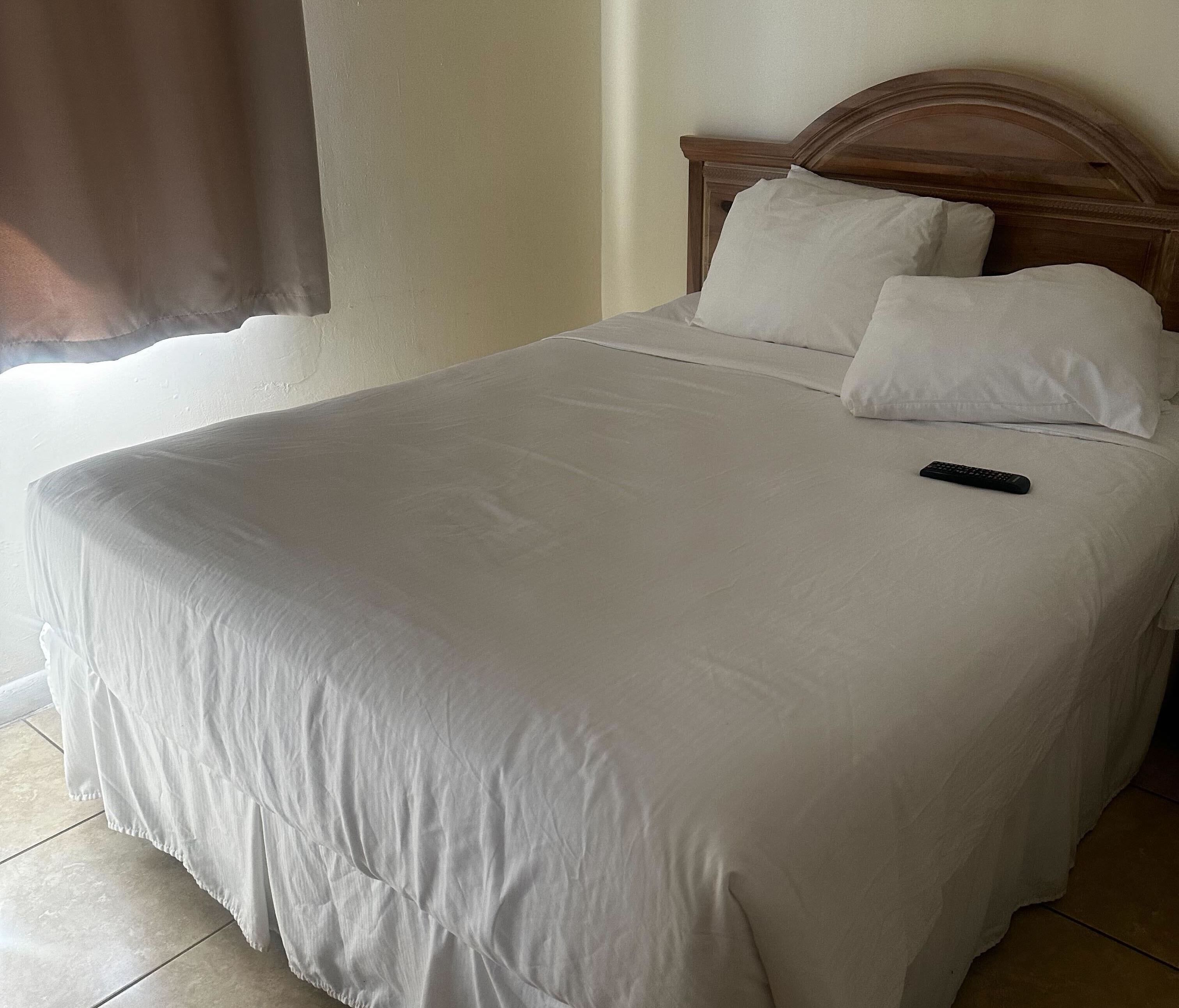 Een netjes opgemaakt bed met witte lakens en kussens, een houten hoofdbord en een afstandsbediening op het bed.