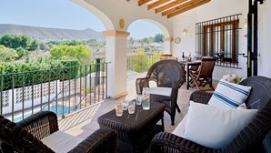 Eine überdachte Terrasse im Freien mit Korbmöbeln, einem Esstisch und Blick auf eine gebirgige Landschaft.