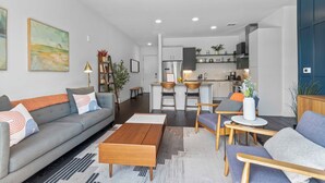 Un salon moderne avec un canapé gris, une table basse en bois et une cuisine en arrière-plan.