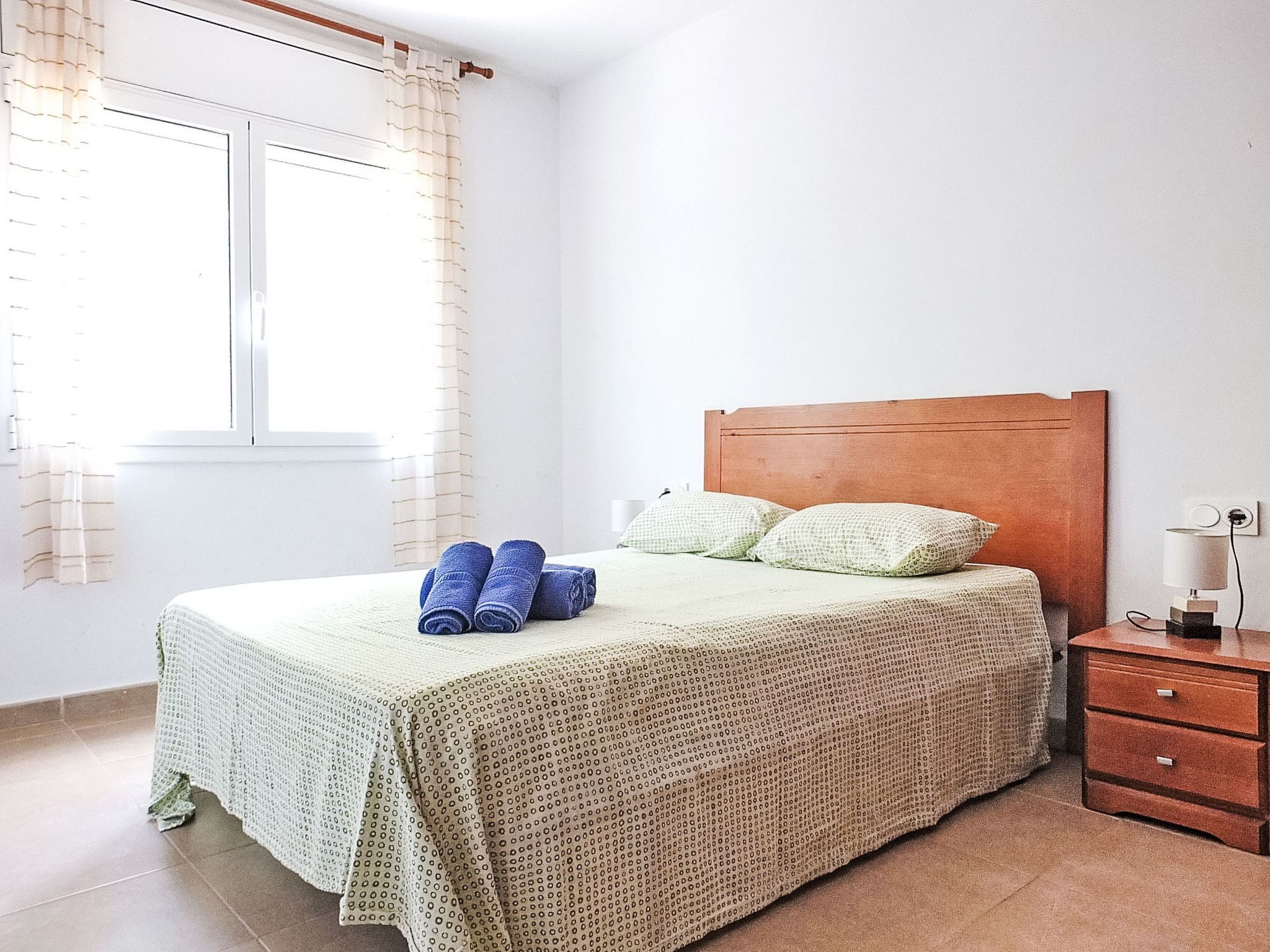 A neatly made bed with a wooden headboard, a bedside table with a lamp, and a window with curtains.