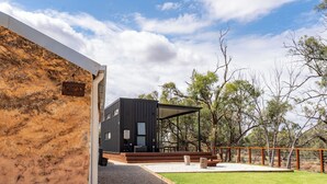 Un edificio moderno de color negro con un área de estar al aire libre cubierta, rodeado por un patio cercado y árboles.